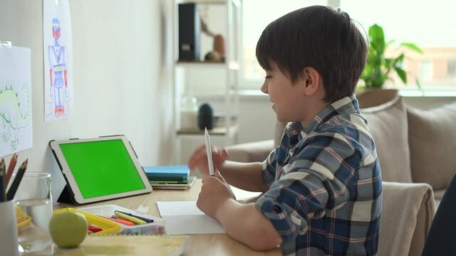 Boy student talk on green screen video class lesson with smile Spbd. Primary school pupil elearning with chroma key tablet, waving hand. Homeschooled child study online with internet teacher tutor