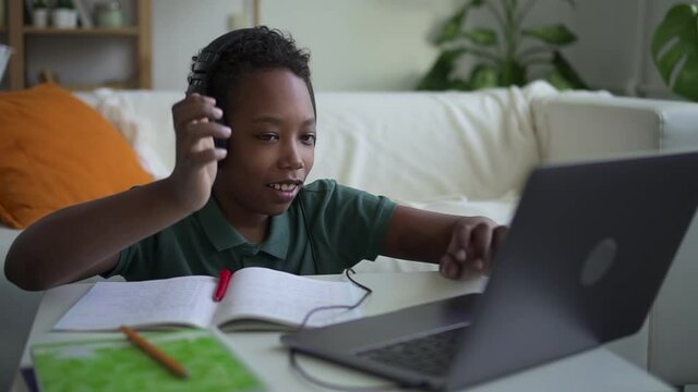 Online primary school education. spbd Happy African-American boy puts on headphones and waves hand to greet teacher and classmates via laptop at home