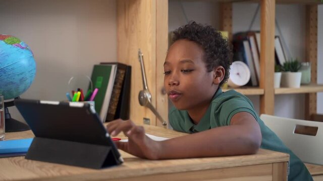 Online education at home. Happy African-American schoolboy does test spbd on tablet sitting at table with copybook and globe close side view - Powered by Adobe