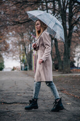 A young, slender, beautiful girl in a coat walks in the rain with a transparent umbrella in the park