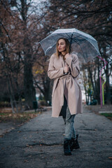 A young, slender, beautiful girl in a coat walks in the rain with a transparent umbrella in the park