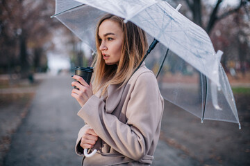 A young, beautiful girl with a disposable cup of coffee walks in the park under a transparent umbrella in the rain