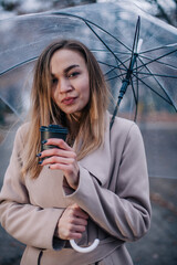 A young, beautiful girl with a disposable cup of coffee walks in the park under a transparent umbrella in the rain