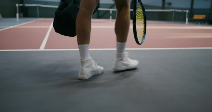 A young man with tennis equipment in his hands is walking on a tennis court