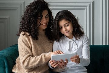 Happy young curly-haired mom sits on the couch in the living room and relaxes with her young daughter, watching a cartoon on a tablet together, smiling mom and schoolgirl have fun playing on the