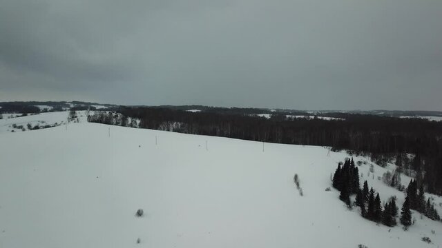 Aerial shot in movement of snowed hill with green spruces on background and dark clouds when it's snowing