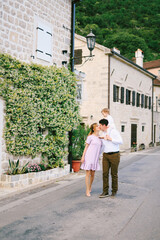 Fototapeta na wymiar Mom kisses dad, holding little daughter on his shoulders. Family walks down the street with ancient buildings and flowering plants