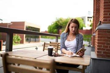 Charming female blogger with modern cellphone gadget and education textbook sitting at cafeteria table and writing informative notes, Caucaisan hipster girl with smartphone studying at sidewalk