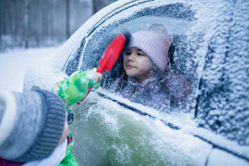 Little boy brushes snow from a car window, into which girl looks out. It looks as if a boy is cleaning a girl, funny moment. Family road trip, active weekend in any weather, winter car safety
