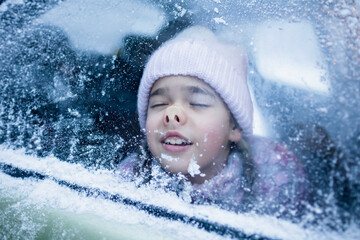 Little girl wearing in winter clothes pressed her face against car window glass funny flattening...