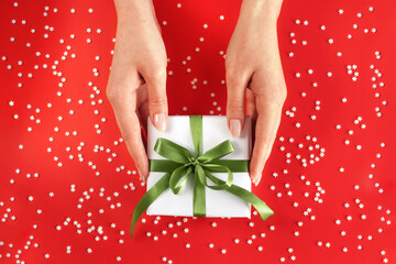 Nice female hands holding gift box wrapped in white paper with green ribbon on festive red background with many snowflakes. Xmas and New Year postcard design. Giving love and warmth, Christmas concept