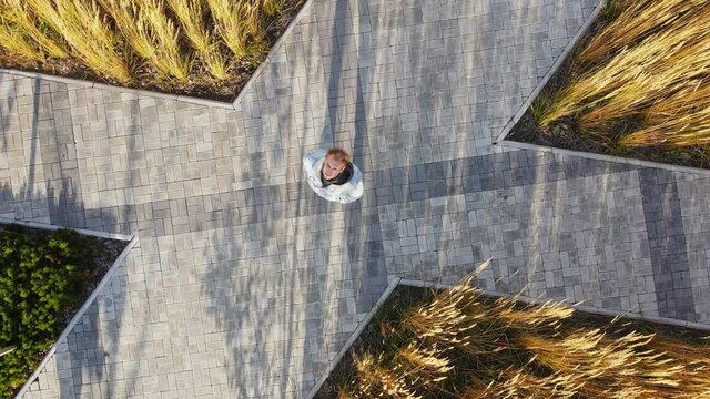 The man waves his hand towards the camera. A man standing against the background of a small park with wheat and street tiles. Shooting from a quadcopter. A small hit on a man from top to bottom