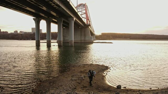 A man alone dances on the sand against the backdrop of a large bridge and the embankment of the river. Dancing in the open air.