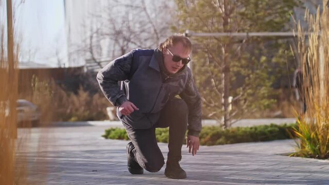 A man dances breakdancing on a street tile in the middle of the city background and wheat growing nearby. A man in a gray jacket is engaged in active sports activities