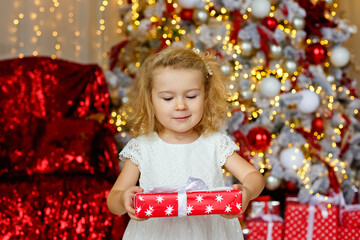 A large portrait of a little curly blonde girl in a white dress with red presents in a room with a Christmas tree and lights of garlands.