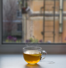A transparent glass cup with hot tea with lemon stands on a white windowsill against the background of the window.