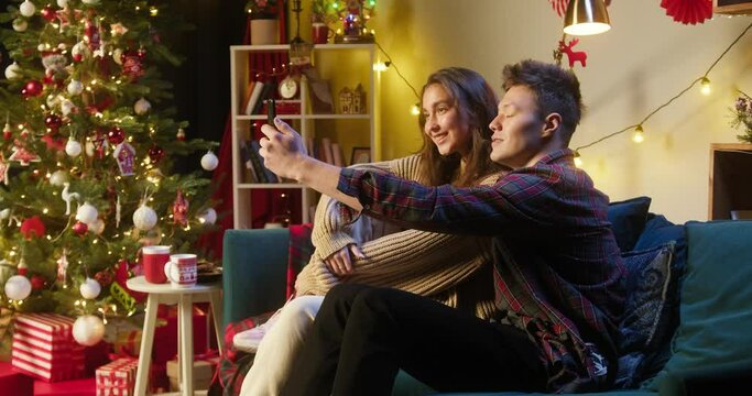 Young couple making selfie on smartphone, sitting on sofa in living room. International friends taking photos on mobile phone. New year evening at home, christmas holidays concept.