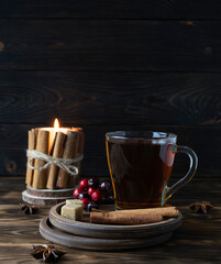 Fragrant hot tea with cinnamon on a wooden saucer. A candle decorated with cinnamon tubes in the background. Berries and pieces of cane sugar on a saucer