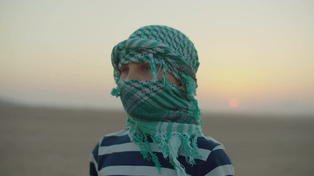 10 years old boy wearing green checkered keffiyeh standing in desert at dawn. Desert sunrise with tourist kid turning to camera.