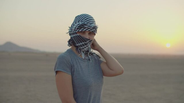 30s woman wearing black and white checkered keffiyeh standing in the desert looking at camera. Caucasian female tourist taking off headdress and smiling at camera. Desert sunrise.