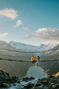 P&auml;rchen sitzt auf einer H&auml;ngebr&uuml;cke hoch in den Bergen, &uuml;ber einem See bei Sonnenaufgang.