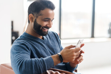 Cheerful hilarious Indian middle-aged man using smartphone sitting on the couch at home, happy eastern guy watching comedy show and laughing, dark-haired hispanic male enjoying online chatting