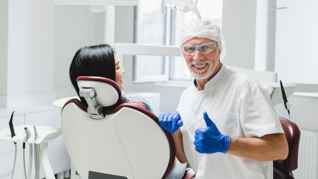 Smiling successful male orthodontist dentist curing operating patient, caries, decay, making veneers, filling teeth looking at camera showing thumb up. Stomatology concept