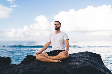 Fototapeta na wymiar Portrait of young male mediate in lotus pose and looking at camera during daytime at coastline rocky stone, calm Caucasian man in casual wear healing during holistic retreat at seashore environment