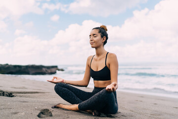 Fototapeta na wymiar Middle Eastern female yogi meditating in lotus pose getting energy from ocean seeking enlightenment and getting mindfulness harmony, calm woman relaxing during concentration workout at coastline