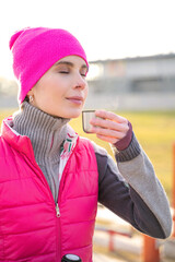 Young beautiful fit woman in warm colorful fitness clothes enjoying smell and drinking hot tea or coffee on workout outdoors on sports ground on frosty early morning sunrise. Holidays, weekend
