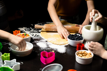 Obraz na płótnie Canvas Children cook cookies together. The shortcrust pastry is rolled out with a rolling pin. Various forms for cutting cookies, nuts, raisins, candied fruits on a dark kitchen table.