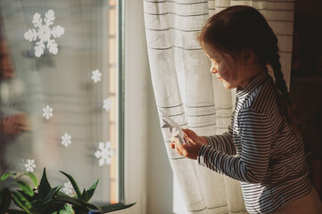 Cute little girl decorates the window of the house with paper snowflakes. Christmas concept