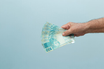 Uma mão de pessoa idosa com algumas notas de 100 reais com fundo azul. Dinheiro brasileiro....
