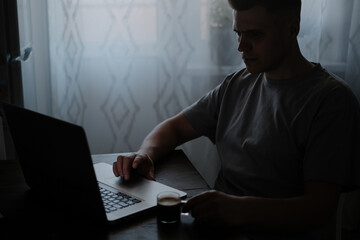 A man in the kitchen at the table in the morning drinks coffee and looks at a laptop