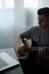 A man at home plays the guitar and looks into a laptop