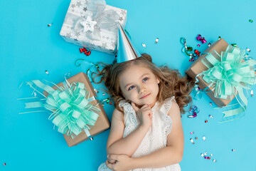 a thoughtful little girl in a cotton white dress and a festive hat on a blue background with gifts and a serpentine makes a wish
