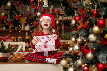 a little cute girl child in a red sweater and a Santa Claus hat in a dark kitchen with a Christmas tree rejoices with gifts and waits for the new year or Christmas