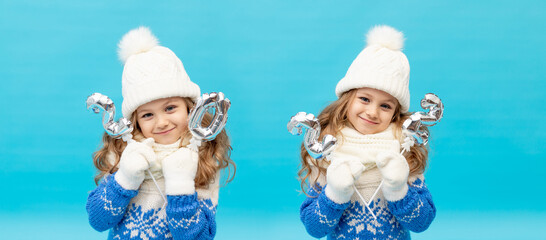 a little girl child on a blue background in the studio in a winter hat and sweater holds the numbers of the year 2022 on a banner, the concept of new year and Christmas, space for text