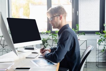 Side view of developer writing on notebook near computers with blank screen and smartphone in office
