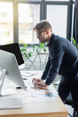 Side view of developer in eyeglasses using computers near smartphone and paper in office