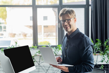 Young programmer in eyeglasses smiling at camera while using laptop near computers in office