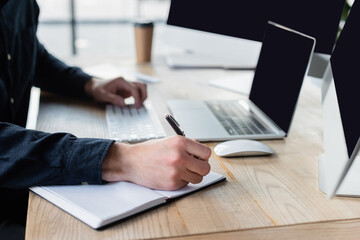 Cropped view of programmer writing on notebook near computers on blurred background