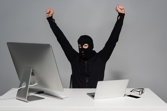 Excited hacker in balaclava showing yes gesture near gadgets on grey background