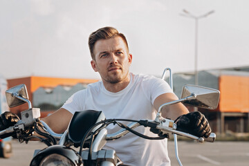 Attractive stylish young man in white T-shirt sits on a motorcycle. Portrait handsome biker posing on a bike. Life style photography. Urban background.