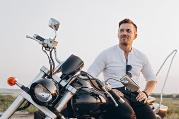 Stylish young attractive guy is sitting on a motorcycle. A nice young man in a white shirt sits and rests on a bike. Hot sunny day. Sky background
