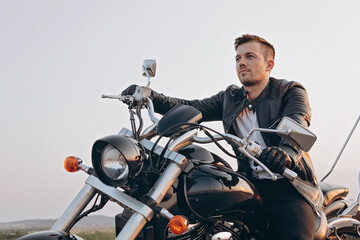 Fototapeta na wymiar Stylish young attractive guy is sitting on a motorcycle. A handsome young man in a black leather jacket is driving a black bike. Sunny evening.
