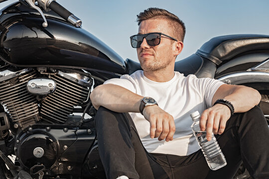 Young attractive guy in sunglasses is holding a bottle of clean water. A nice young man in a white T-shirt sits and rests on the background of a motorcycle. Hot sunny day. Life style