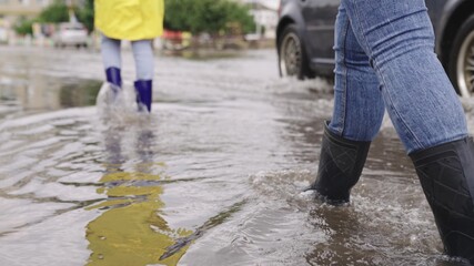 girls in raincoats and rubber boots walk along road flooded with torrential rains, their feet walk through puddles city, splashing water to the sides, the flood is on street, car is driving on water - obrazy, fototapety, plakaty