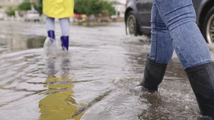 Foto op Aluminium girls in raincoats and rubber boots walk along road flooded with torrential rains, their feet walk through puddles city, splashing water to the sides, the flood is on street, car is driving on water © Валерий Зотьев
