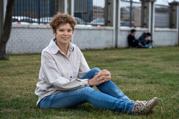 Obraz premium Portrait of a smiling european girl with short brown curly hair wearing beige corduroy shirt sitting on the green grass on the lawn in the park 